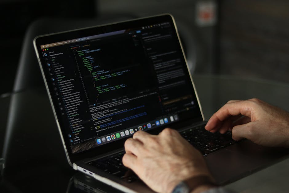 A close-up view of a person's hand typing on a laptop keyboard, with visible code on the screen. The laptop is placed on a wooden surface, possibly a desk, and the person's wrist is adorned with a wri